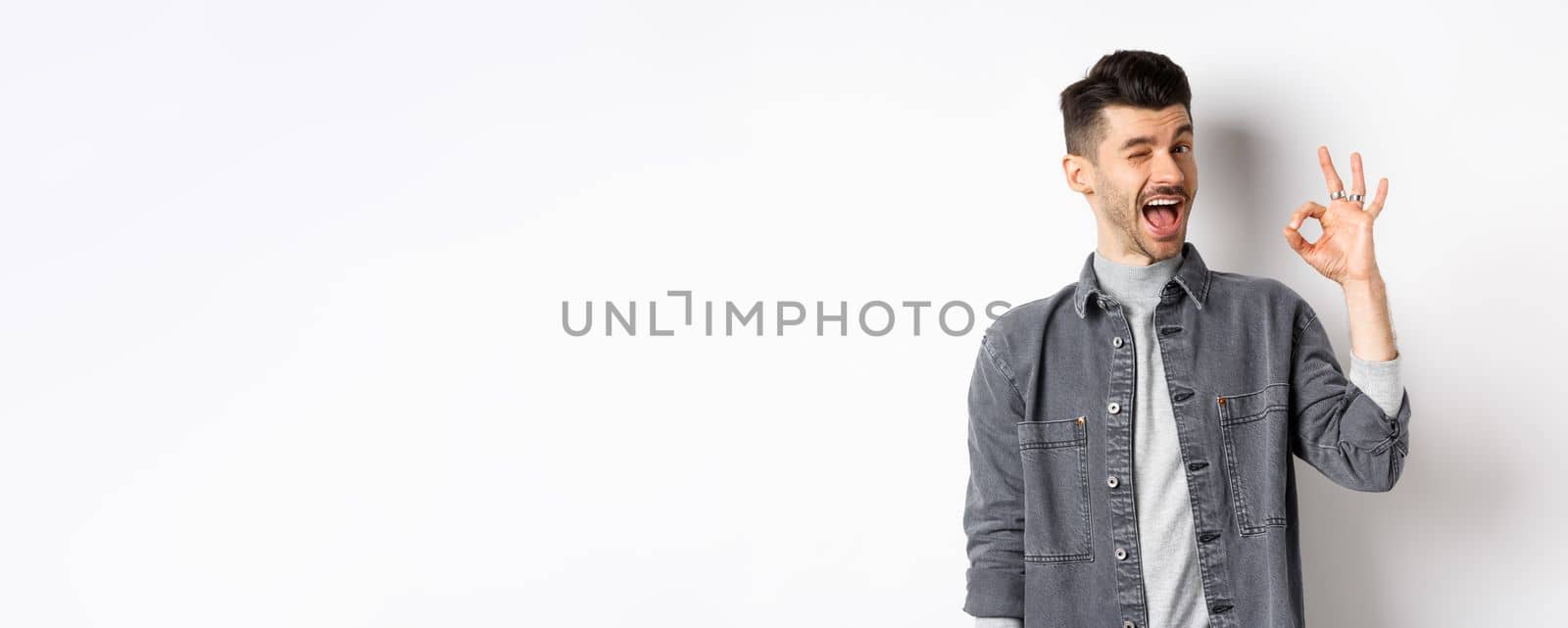 Excited handsome guy with moustache, winking and showing okay sign, smiling pleased, assure all good, praise nice work, making good job gesture, standing against white background.