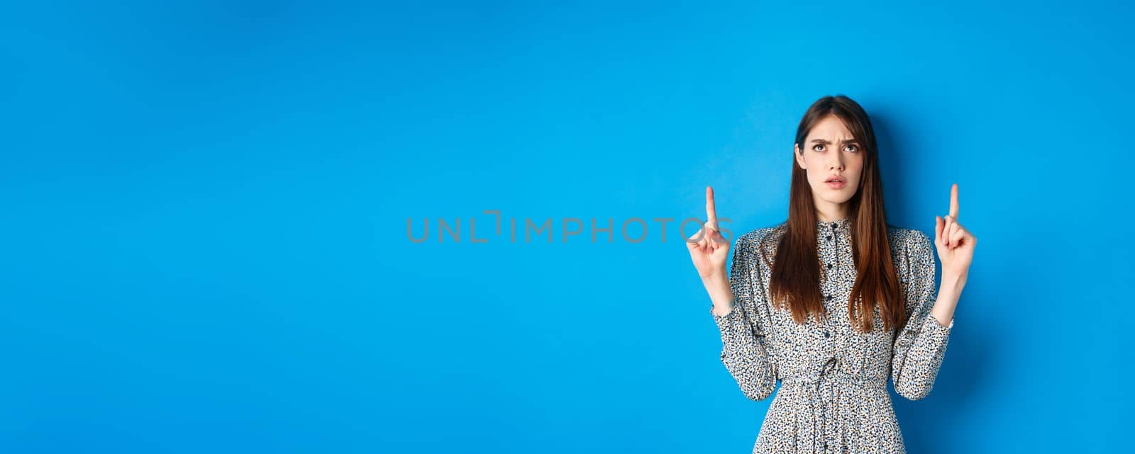 Confused attractive woman in dress frowning, pointing fingers up and looking puzzled, cant understand something, stare at strange thing, blue background.