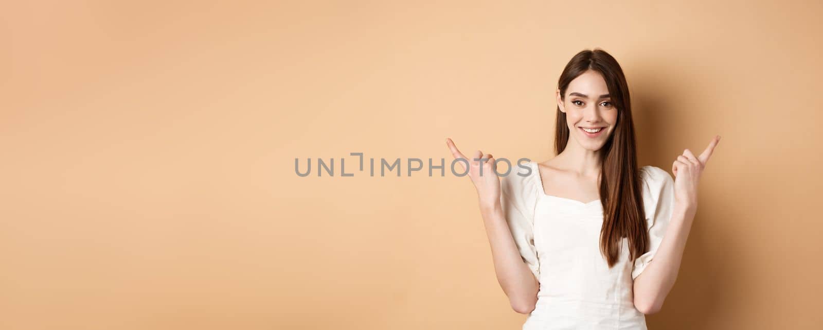 Beautiful young woman in dress pointing fingers sideways, show two ways, giving choice and smiling, standing on beige background.