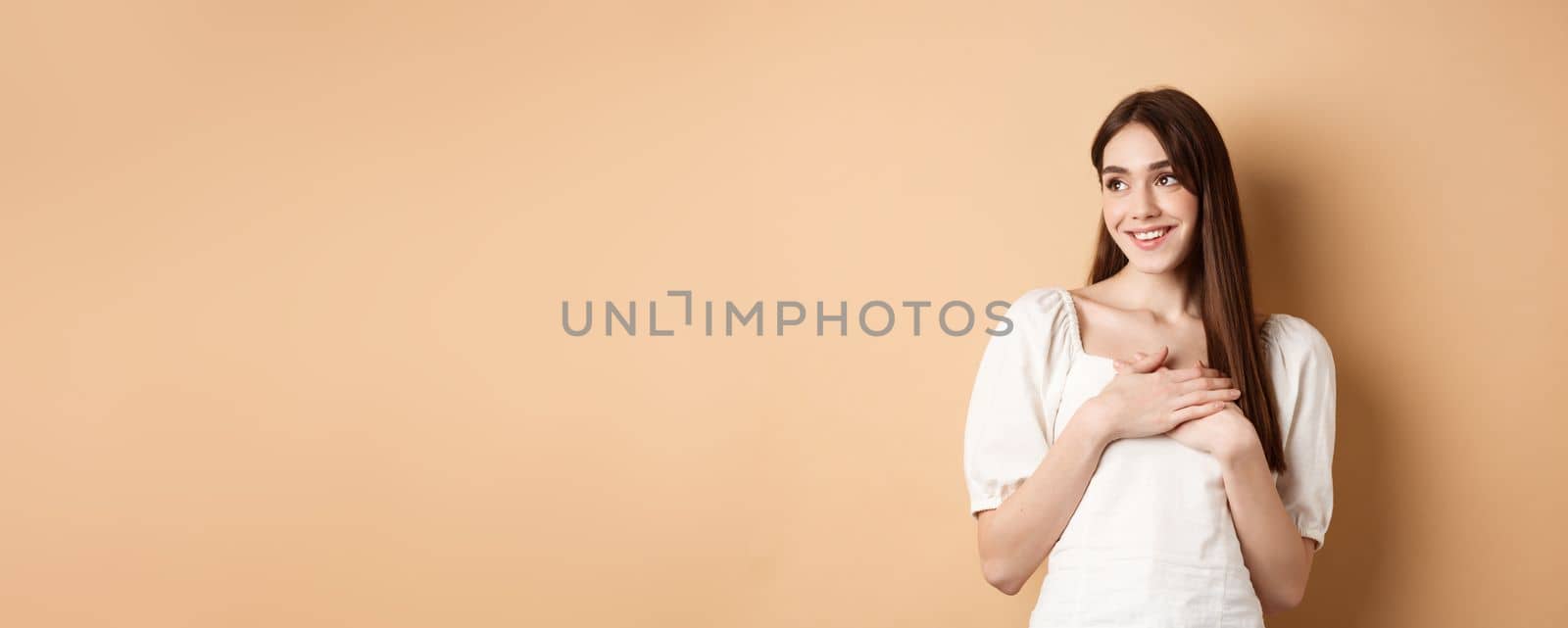 Romantic young girl in dress holding hands on heart, smiling and looking at empty space thankful, feeling gratitude, standing on beige background.