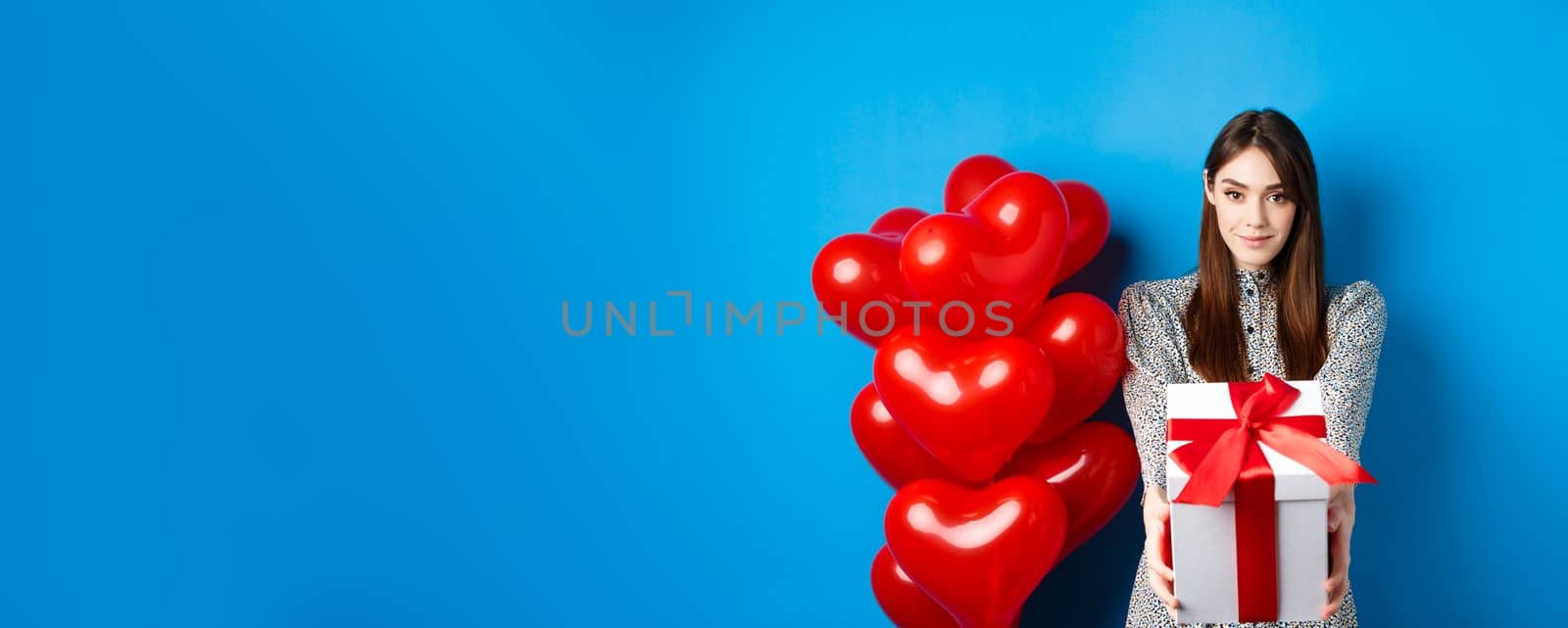 Valentines day. Romantic and cute girl stretching out hands with present, giving gift box to lover and smiling, standing near hearts balloons, blue background.