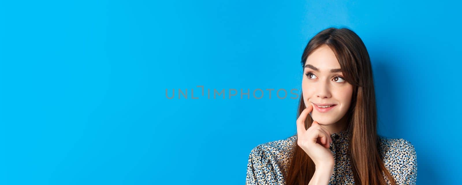 Interesting. Pensive young girl looking aside at logo and smiling, touching lip thoughtful, have an idea, standing on blue background.