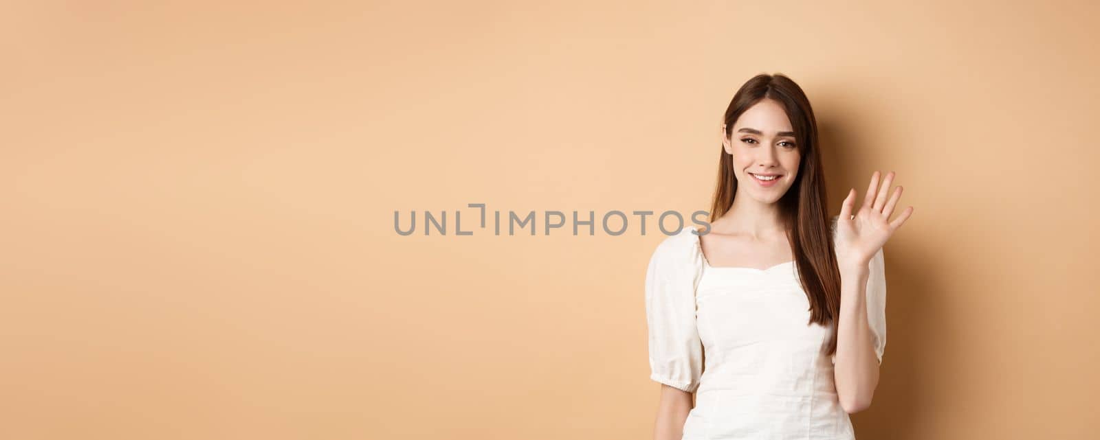 Friendly smiling woman say hello, waiving hand to greet you, standing cheerful on beige background by Benzoix