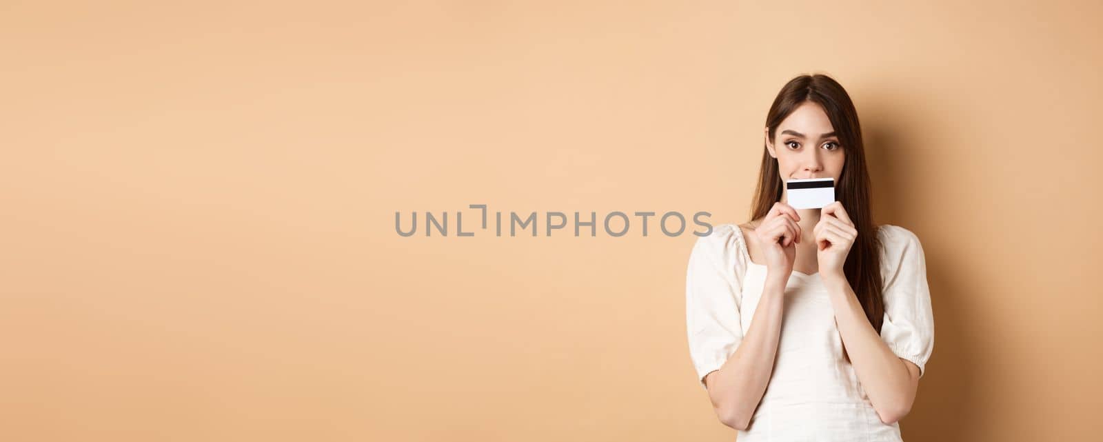 Excited girl holding plastic credit card on lips and smiling, thinking of shopping, going to buy something, standing on beige background.