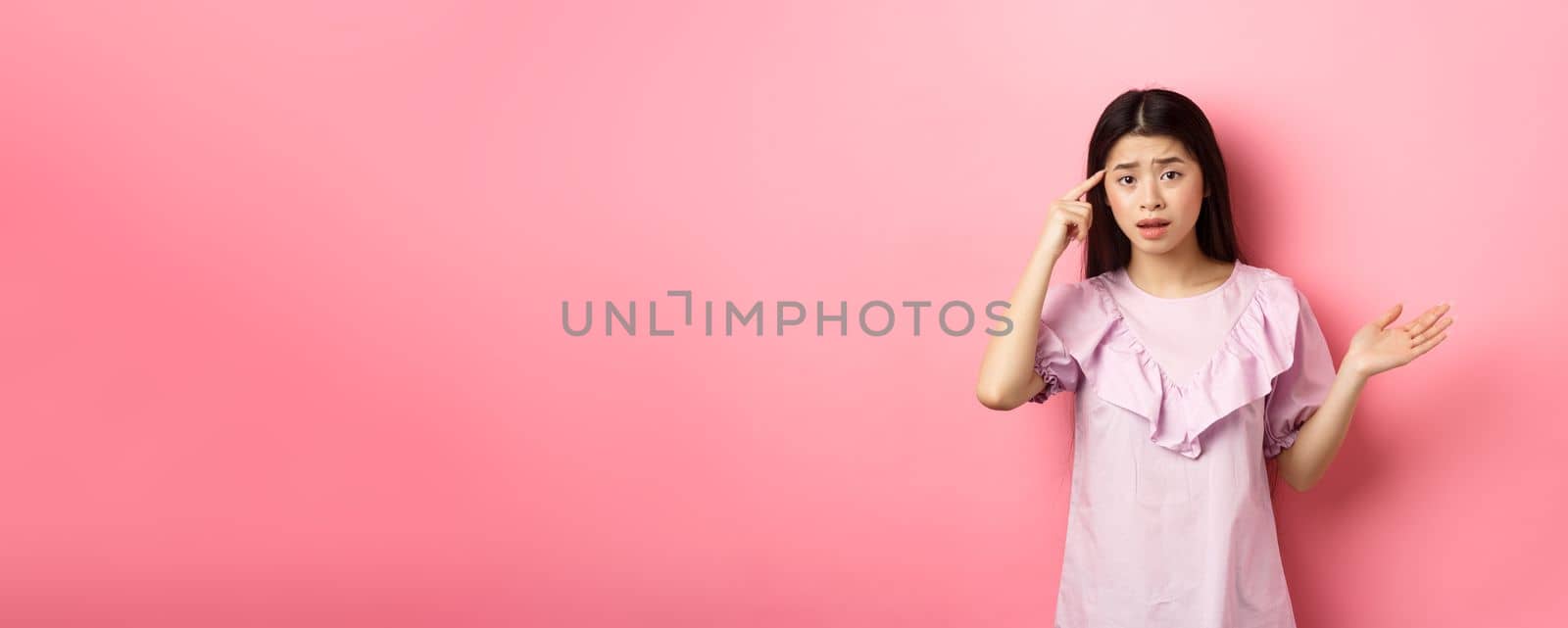 Are you crazy. Shocked asian woman pointing at head temple and complaining someone stupid action, scolding person, standing against pink background.