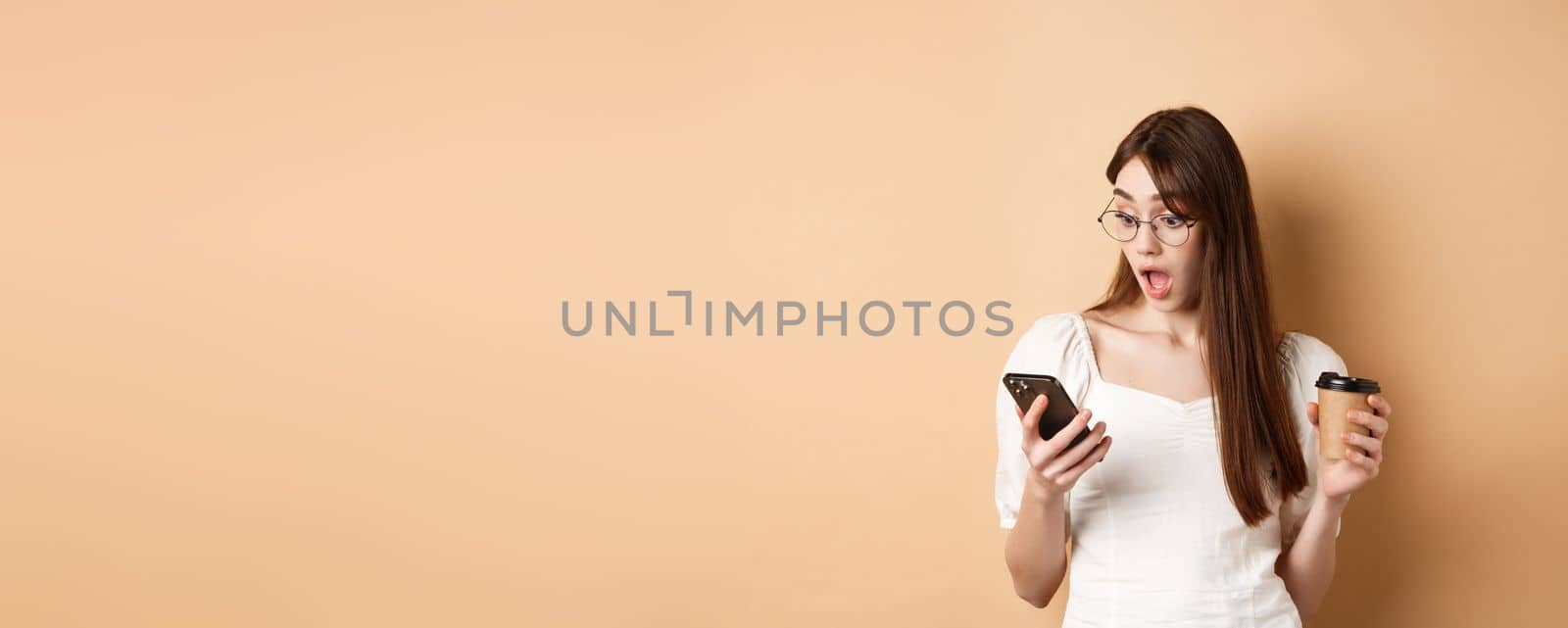 Surprised girl reading exciting news on cell phone, drop jaw amazed, standing with coffee cup on beige background.