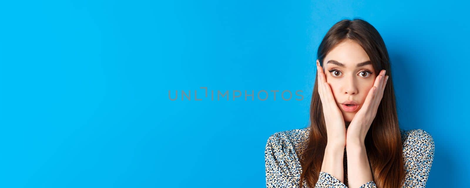 Close-up portrait of surprised cute woman holding hands on cheeks and gasping, stare at camera shocked, standing against blue background by Benzoix