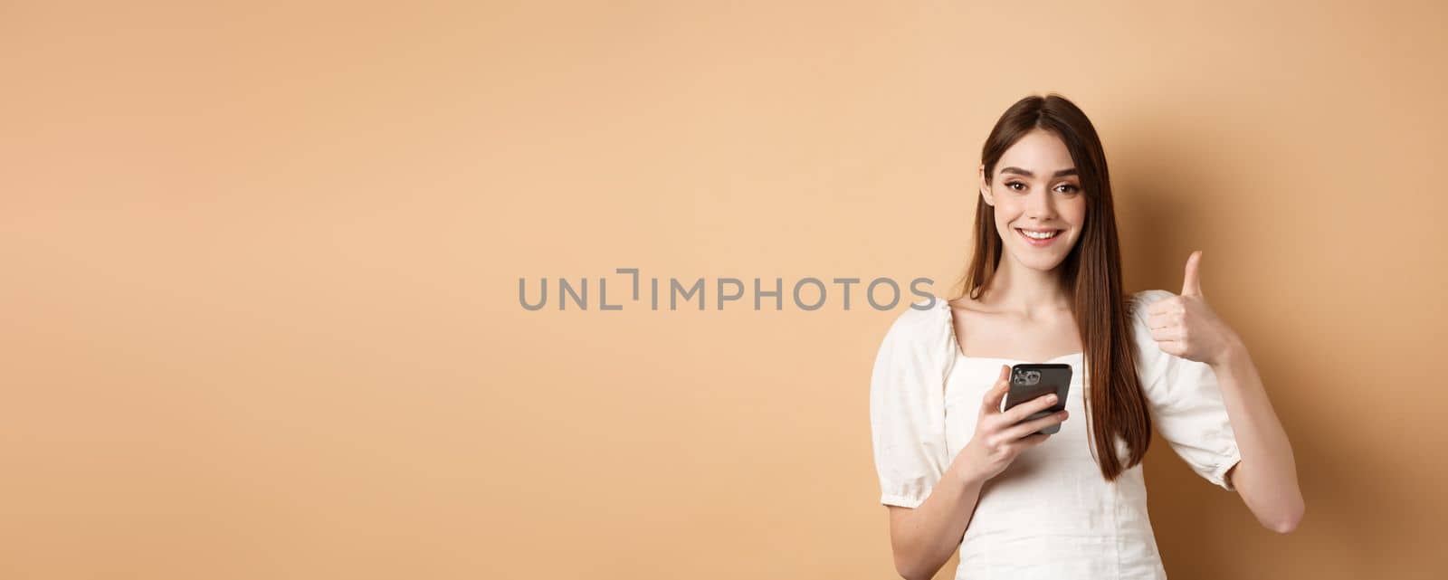 Online shopping concept. Smiling happy woman showing thumb up after using smartphone, holding phone and say yes, standing on beige background by Benzoix