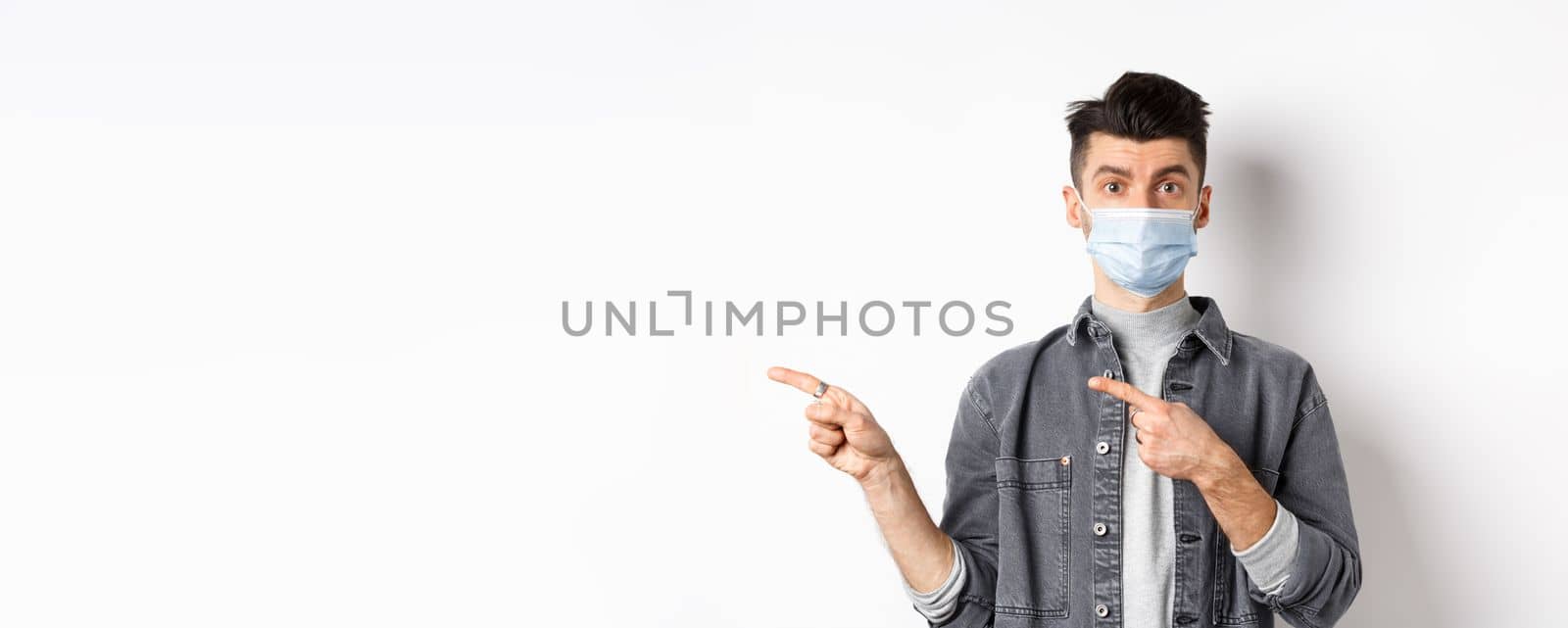 Pandemic lifestyle, healthcare and medicine concept. Young guy in face mask inviting look here, pointing fingers left at logo, standing on white background.