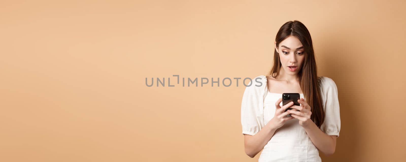 Woman look surprised at smartphone screen, reading shocking message on cell phone, standing amazed on beige background by Benzoix