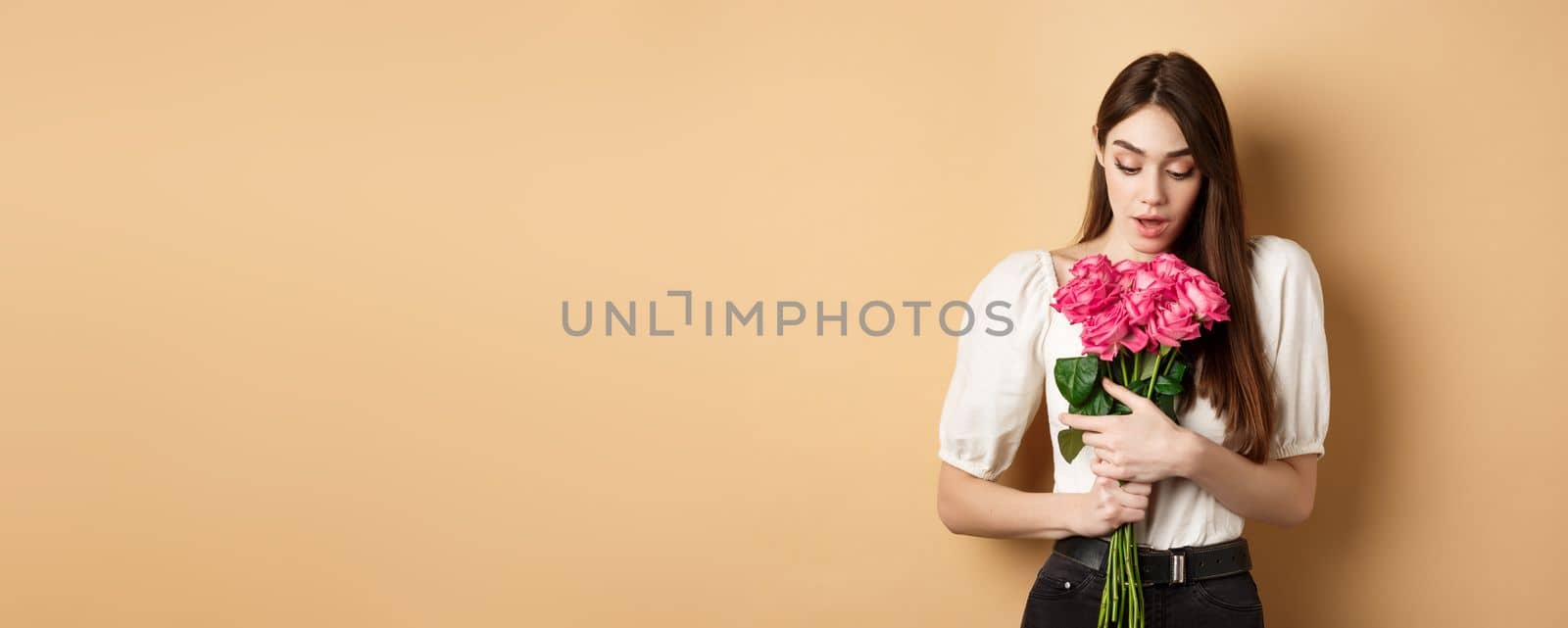 Valentines day. Surprised tender girl looking at beautiful pink roses, receive romantic gift from lover, standing on beige background.