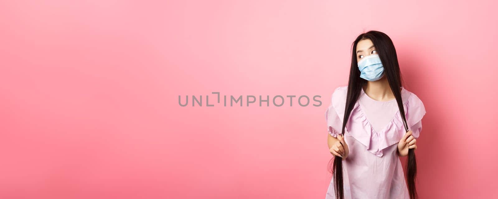 Healthy people and covid-19 pandemic concept. Stylish japanese girl in medical mask posing, looking aside and playing with long hair, standing against pink background by Benzoix