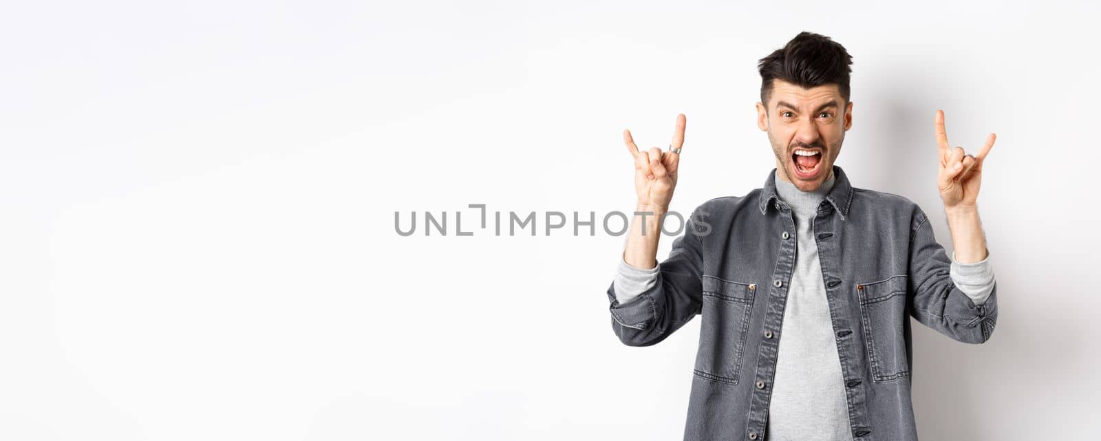 Rock on. Sassy young man shouting and showing heavy metal horns sign, attend awesome concert, having fun, standing on white background.