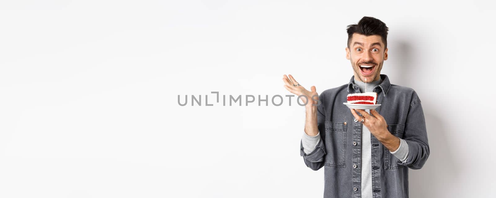 Excited man celebrating birthday, looking happy, holding cake with candle, making wish, standing happy against white background.