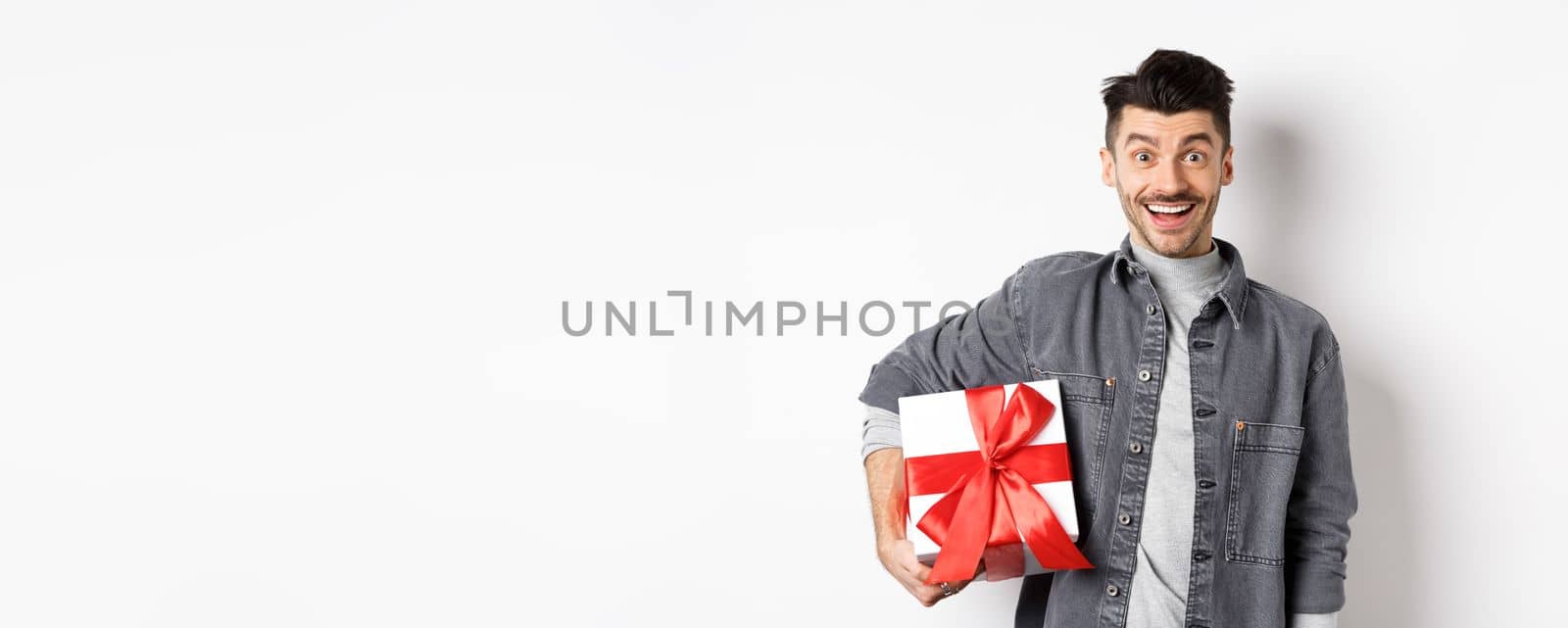 Happy Valentines day. Surprised and happy guy holding gift box and look at camera, smiling amazed, celebrating holiday, bring present on romantic date, white background.