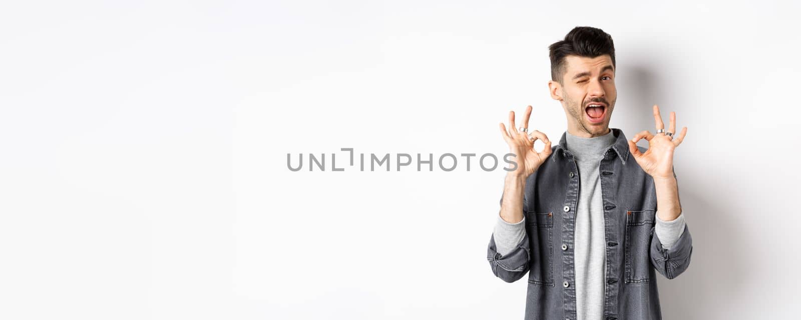 All okay. Handsome caucasian man showing OK signs and smiling, winking to praise good thing, recommending excellent offer, standing on white background by Benzoix