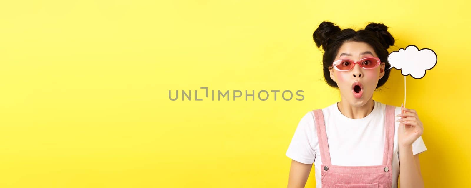 Summer and fashion concept. Excited party girl in sunglasses, holding comment cloud party mask and gasping amazed, standing impressed on yellow background.