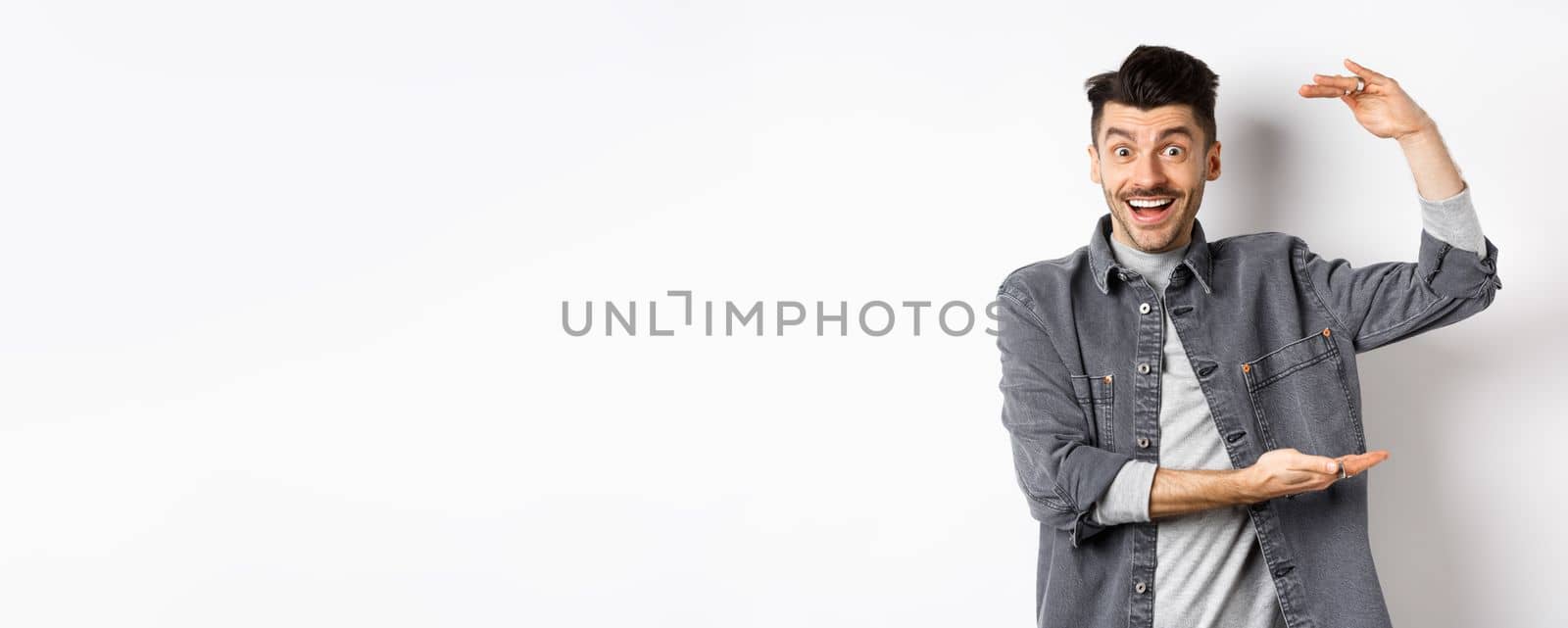 Cheerful guy smiling and showing big size, showing large thing and looking excited, standing against white background by Benzoix