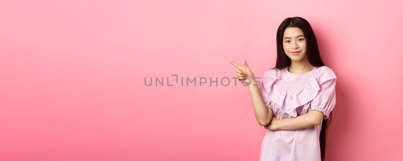 Beautiful teenage asian girl pointing finger left, smiling confident, showing advertisement on pink background by Benzoix