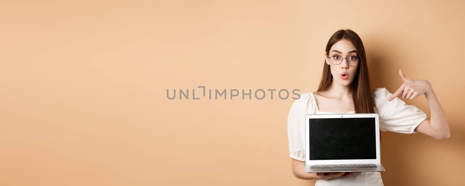 Excited girl in glasses making presentation on computer, pointing hand at laptop screen and say wow, standing on beige background.