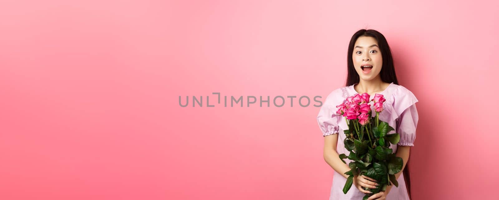 Korean teen girl in dress having romantic date on Valentines day, holding bouquet of roses and looking surprised at camera, receive gift on date from lover, pink background by Benzoix
