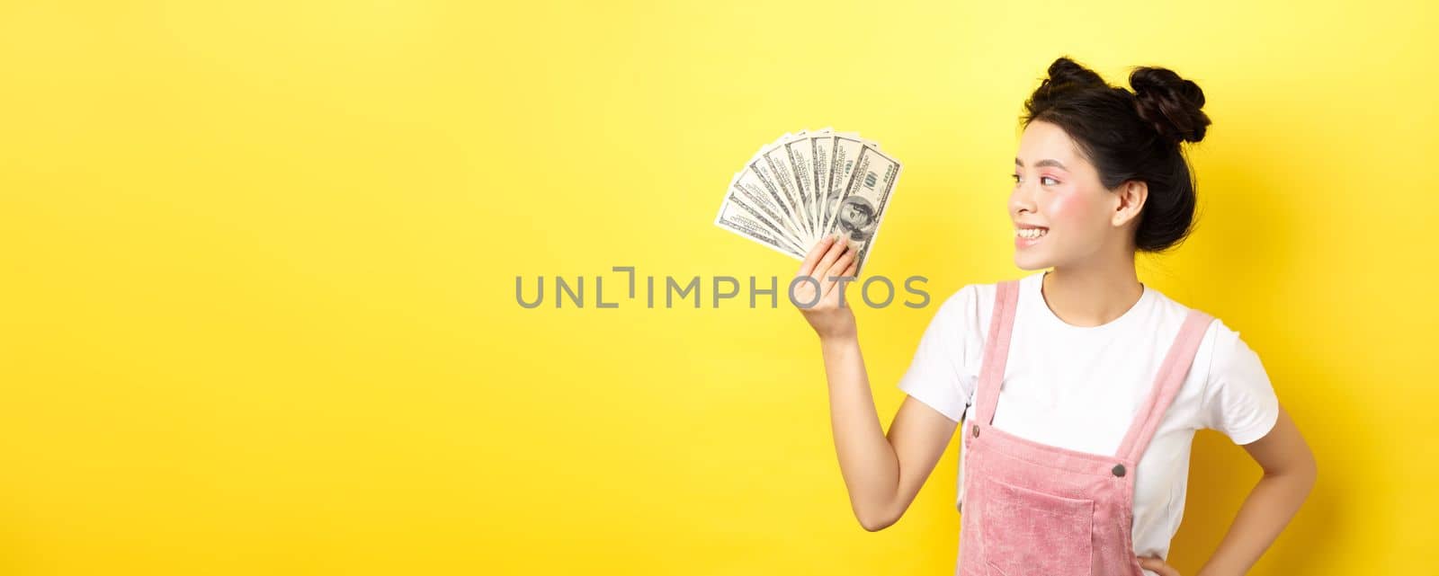 Shopping. Rich and stylish asian female model showing money, looking at dollar bills with pleased smile, standing on yellow background.