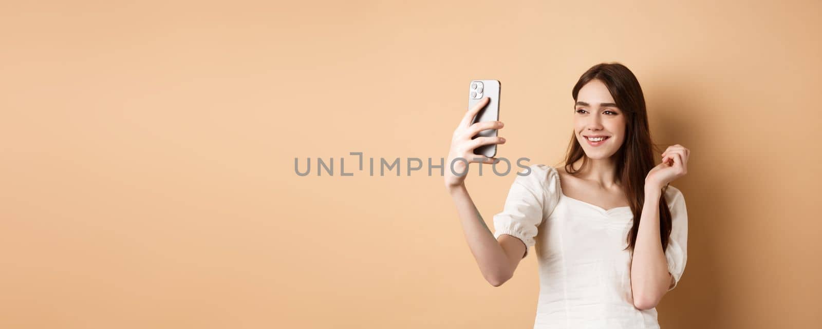 Beautiful woman taking selfie on smartphone, posing for phone photo, standing on beige background by Benzoix