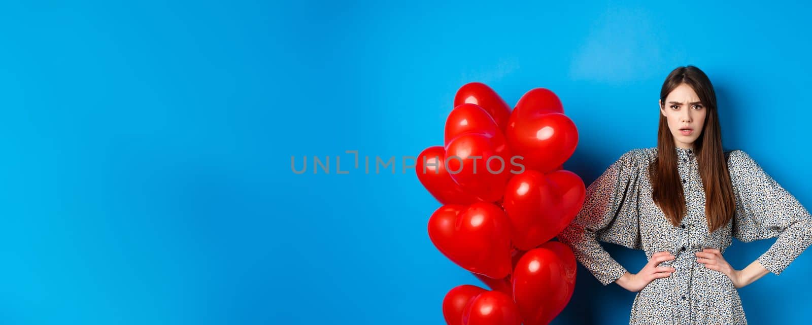 Valentines day. Angry and confused girlfriend in dress, standing near red heart balloons and frowning annoyed at camera, standing near blue background by Benzoix