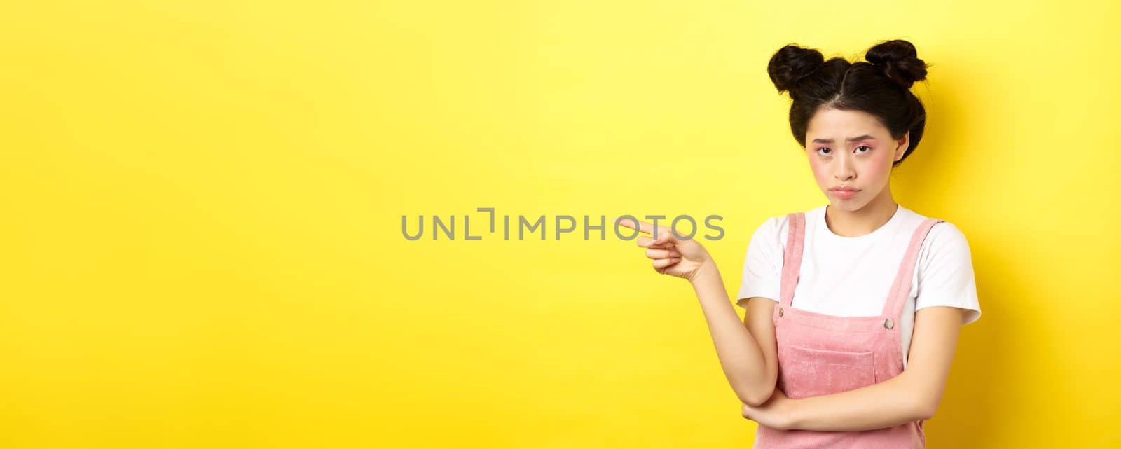 Sad and gloomy asian girl with glamour makeup, frowning unfair and pointing left at logo, looking disappointed, standing on yellow background.