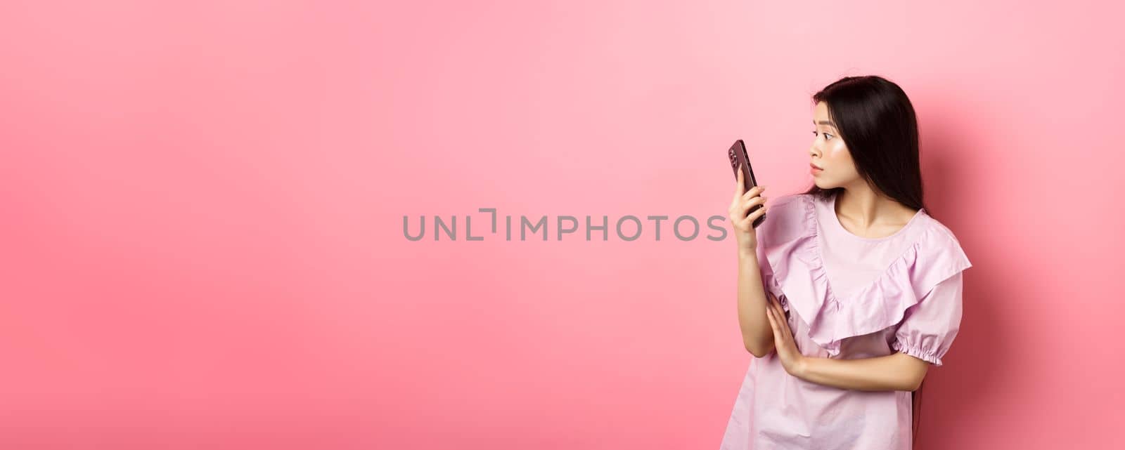 Asian girl look surprised at smartphone screen, reading message, standing in dress against pink background.