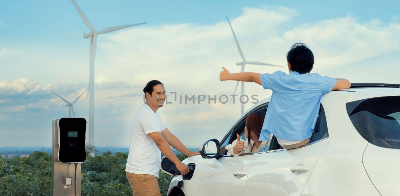 Concept of progressive happy family enjoying their time at wind farm with electric vehicle. Electric vehicle driven by clean renewable energy from wind turbine generator for charging station.