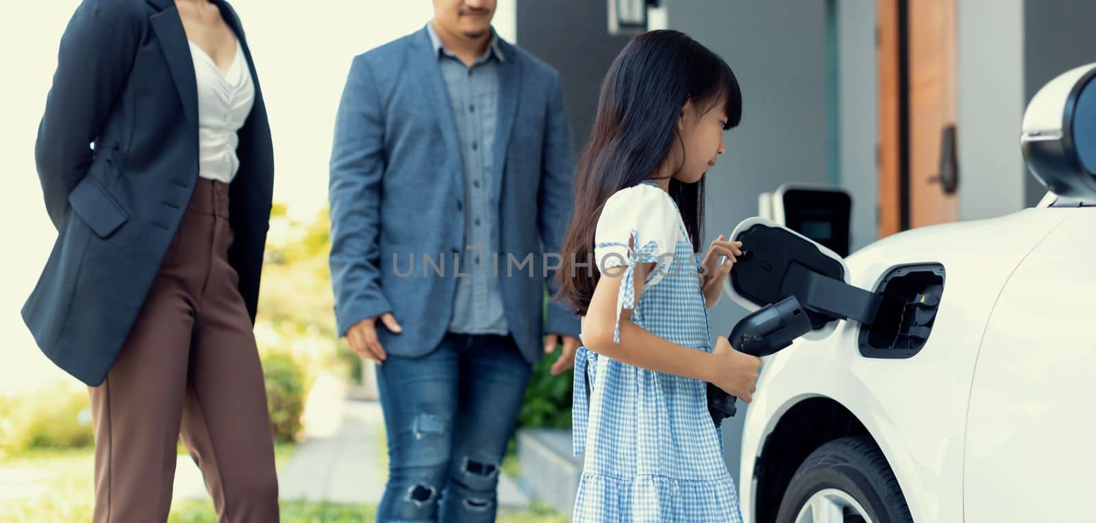 Progressive young parents and daughter with electric vehicle and home charging station. Green and clean energy from electric vehicles for healthy environment. Eco power from renewable source at home.