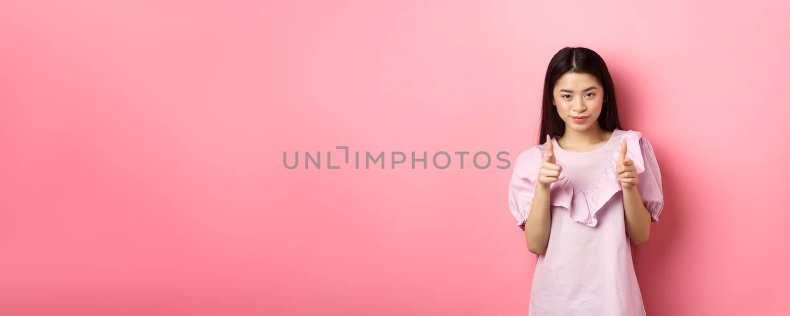 Sassy asian woman pointing at camera, smiling and inviting you, beckon or praise person, standing on pink background by Benzoix