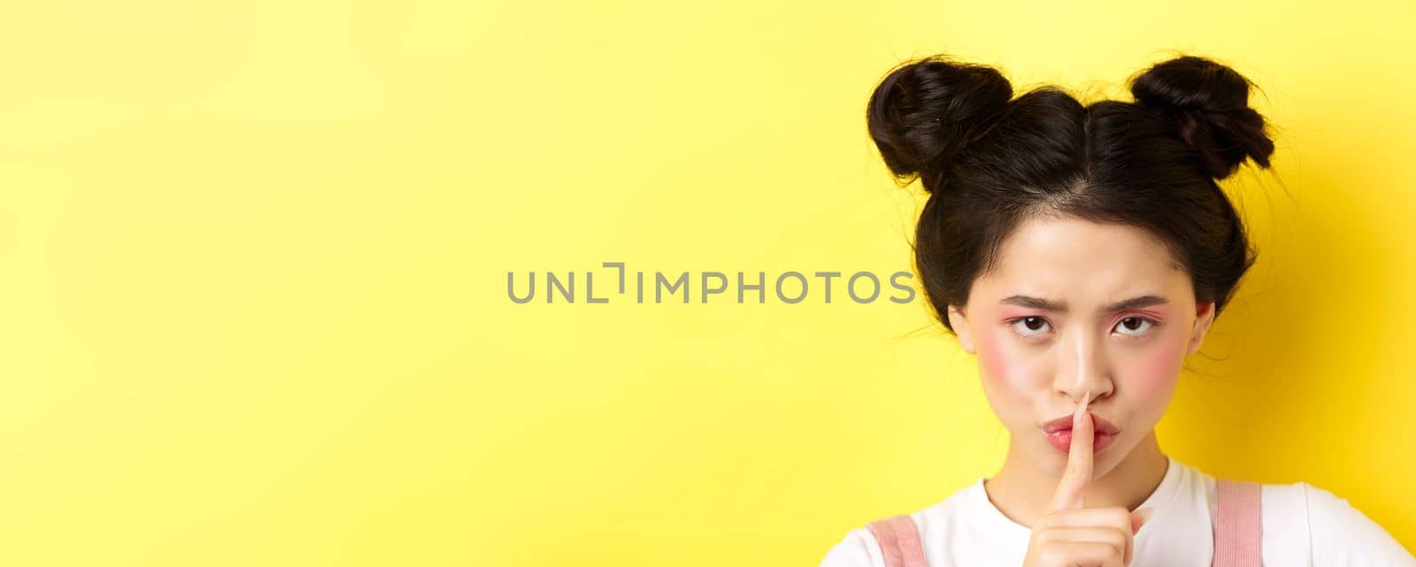 Close up portrait of angry asian girl shushing, press fingers on lips and tell be quiet, scolding loud person, need silence, telling big secret, standing on yellow background.