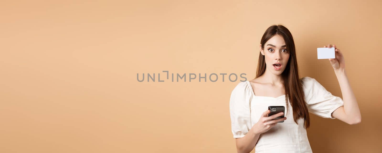 Excited woman showing plastic credit card and using mobile phone app, drop jaw and gasping amazed, checking out bank offer, beige background.