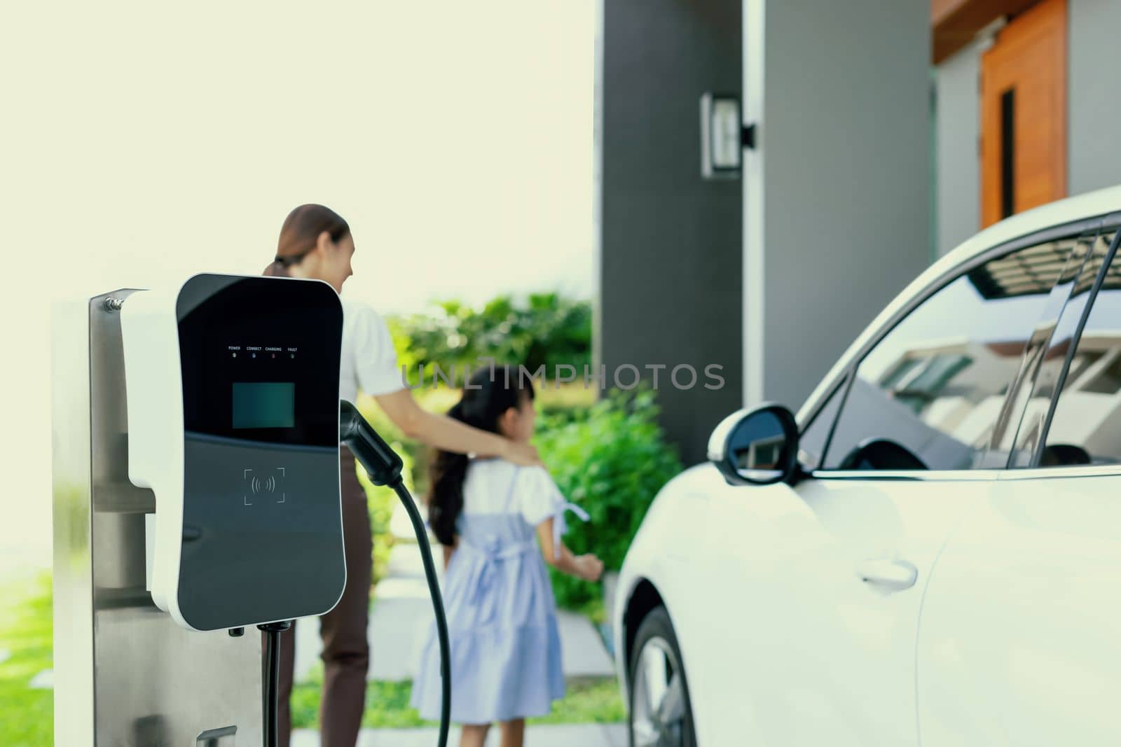 Focus electric charging station power by clean energy for electric vehicle at home with blurred progressive woman and girl walking in background. Home charging station for electric engine car concept
