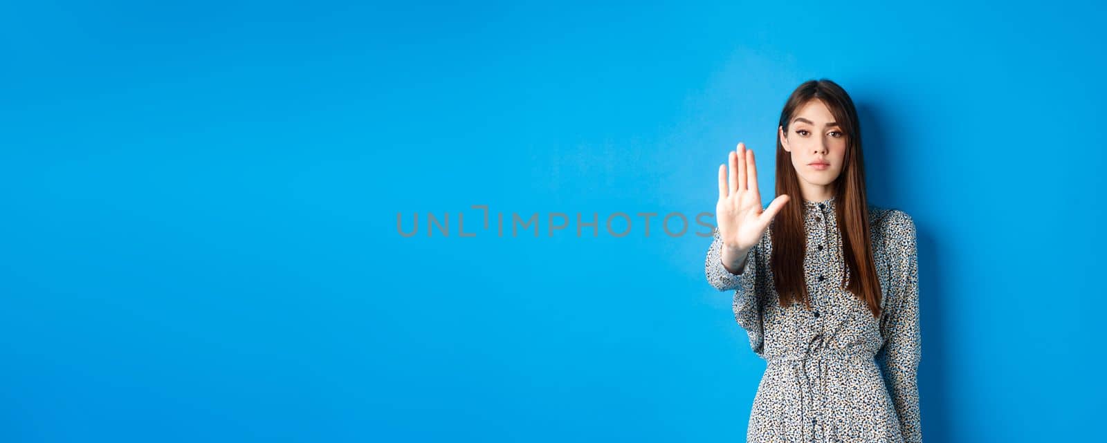 Serious and confident woman with long hair and dress, stretch out hand and say no, make stop gesture, prohibit bad action, standing on blue background.