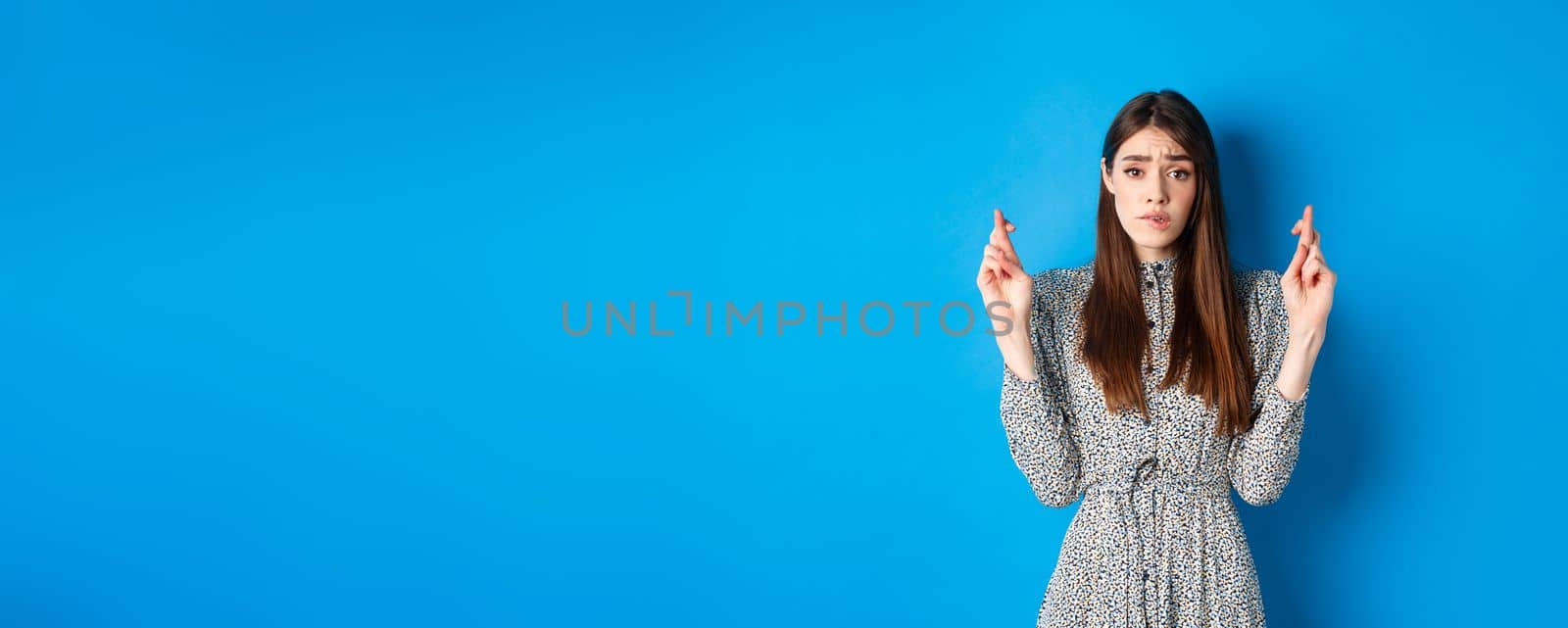 Nervous young woman in dress cross fingers and biting lip worried, praying or making wish, standing on blue background by Benzoix