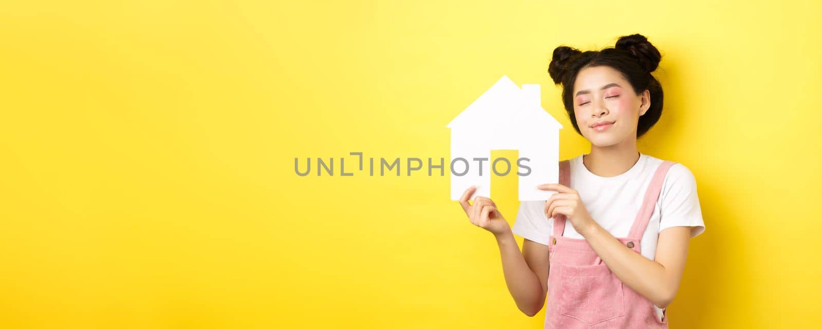 Real estate and family concept. Dreamy smiling asian woman with bright makeup, showing paper house cutout with closed eyes, daydreaming about buying property, yellow background.