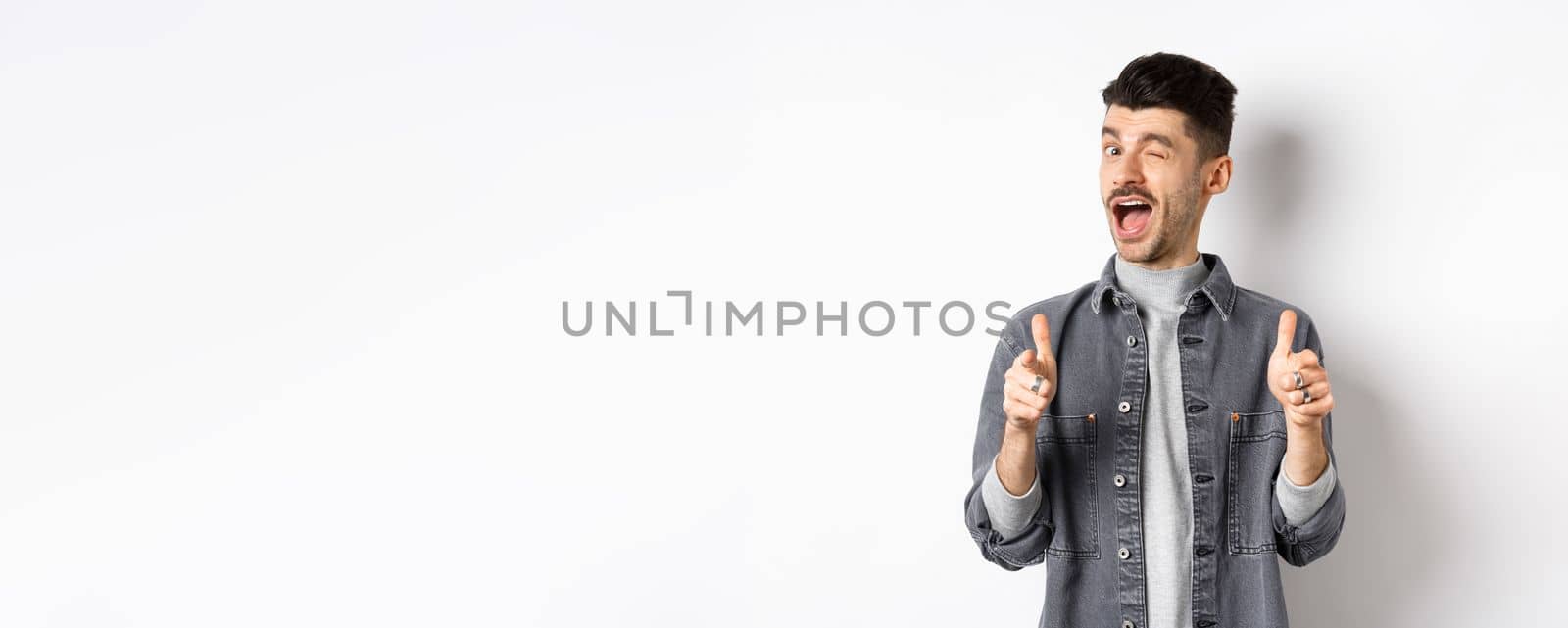 Funny handsome guy winking and pointing fingers at camera, inviting you check out promo, standing on white background.