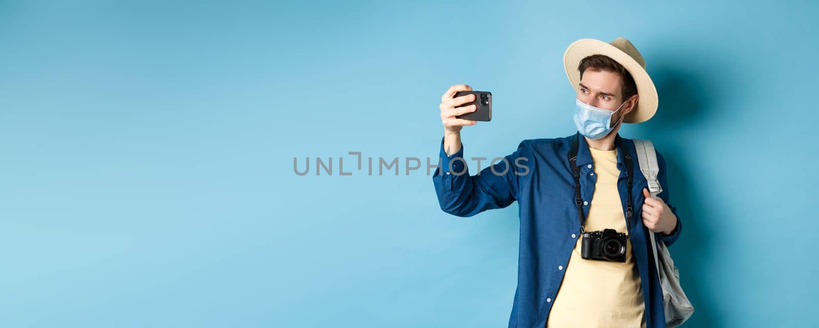 Covid-19, pandemic and travel concept. Male tourist in straw hat and medical mask recording video on smartphone during vacation, taking pics on summer holiday, blue background by Benzoix