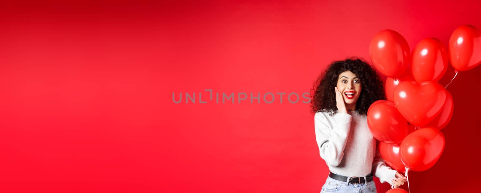 Valentines day. Surprised curly-haired girl holding heart balloons and looking amazed at camera, receive surprise gift on date from lover, standing on red background by Benzoix