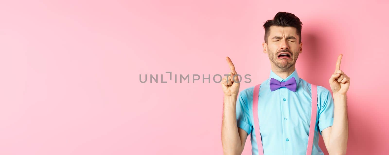 Sad and miserable guy sobbing and crying, pointing fingers up and something disappointed, standing upset on pink background.