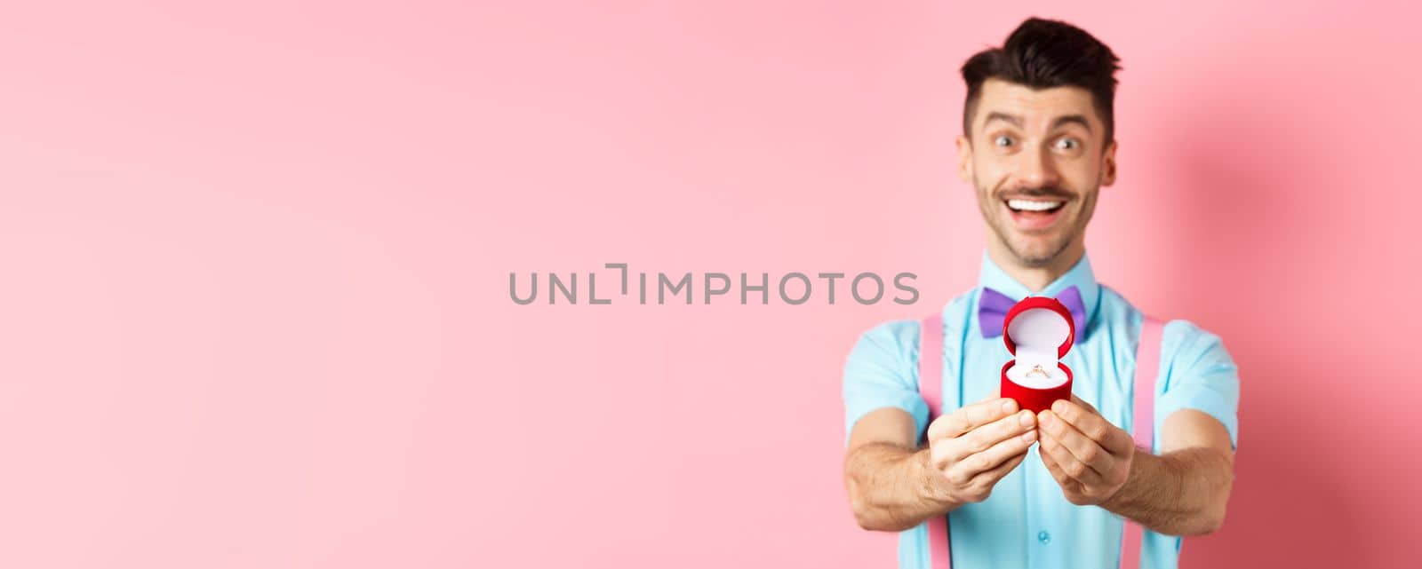 Valentines day. Romantic young man in bow-tie stretch hands with engagement ring and smiling, asking to marry him, making proposal, standing over pink background by Benzoix
