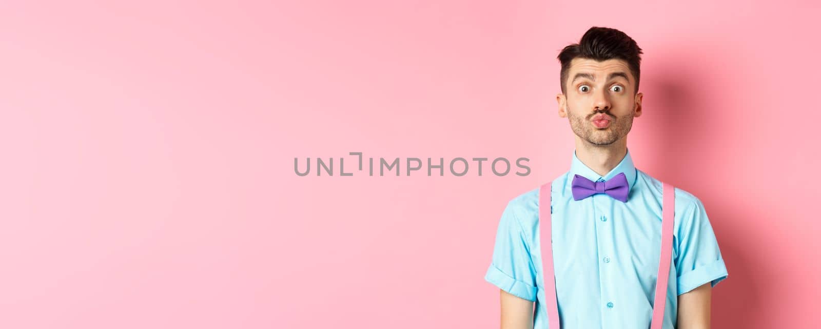 Funny young man waiting for kiss with puckered lips and silly face, standing on romantic pink background. Concept of love and Valentines day by Benzoix