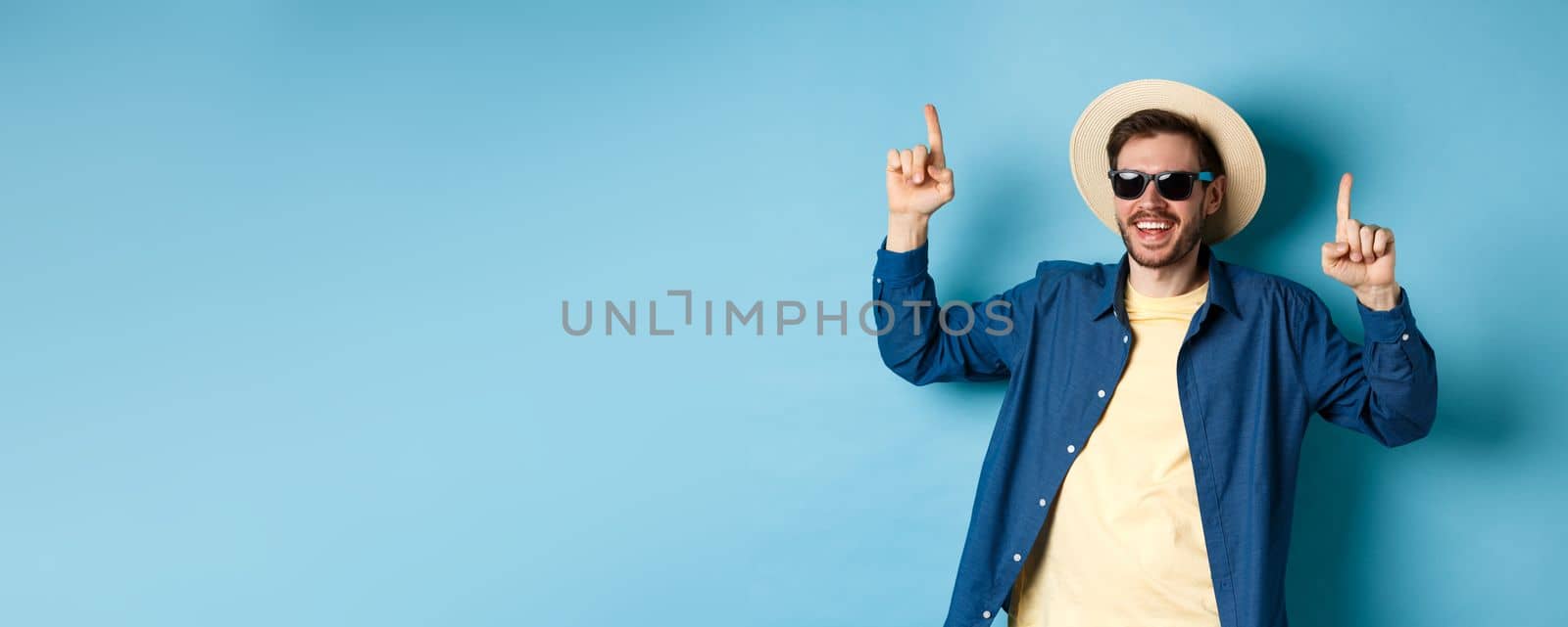 Cheerful caucasian guy in straw hat and sunglasses, dancing and having fun on vacation, standing over blue background. Concept of summer tourism.