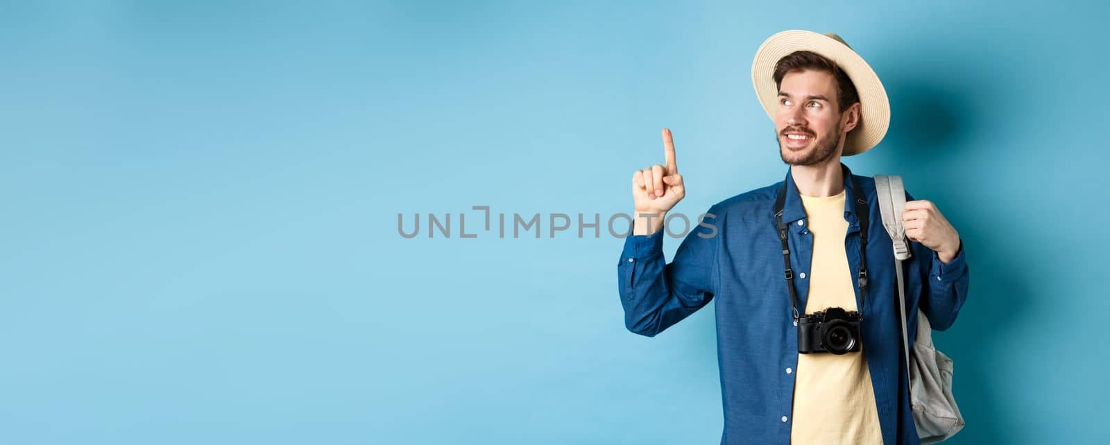 Handsome positive guy on vacation, wearing summer hat, looking and pointing up at logo, smiling pleased, standing with camera and backpack on blue background by Benzoix