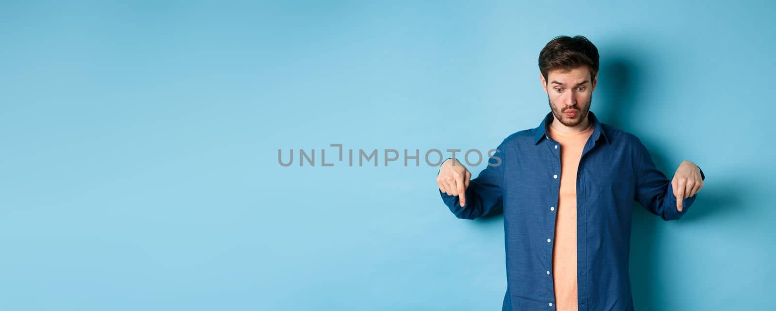 Image of shocked young man in casual clothes, looking and pointing down at something strange, standing on blue background.