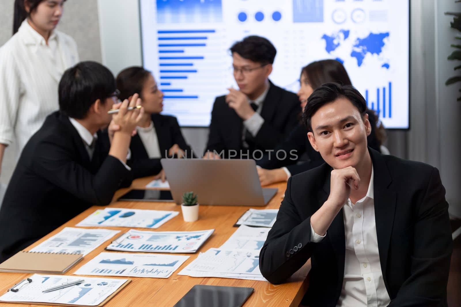 Focus portrait of successful confident male manager or executive in business wear with blurred background of businesspeople, colleagues working with financial report papers in office of harmony.