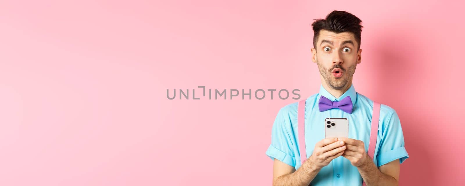 Online shopping. Intrigued guy in bow-tie, checking out internet promo offer on mobile phone, say wow at camera, holding smartphone, standing on pink background by Benzoix
